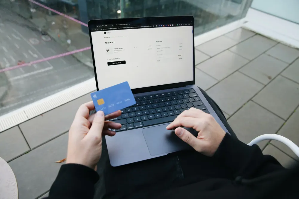 Dizüstü bilgisayarın başında elinde banka kartı tutan biri; ekranda sepet tutarının göründüğü bir online ödeme sayfası açık.