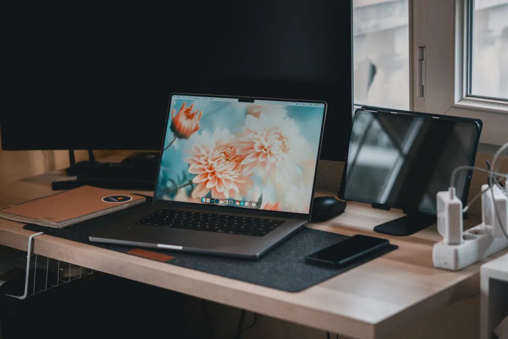 Ahşap bir masada açık duran dizüstü bilgisayar; yanında bir monitör, tablet, telefon ve defter, arkadaki pencereden gelen ışıkla aydınlanıyor.